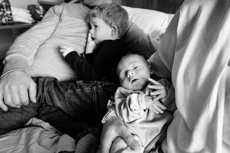 Black and white. Family sits on hospital bed, Son lays on fathers lap while mother holds newborn baby