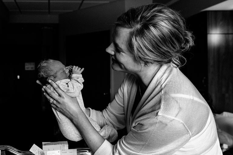 Black and white, Mother smiles while picking up sleeping newborn baby in the hospital.
