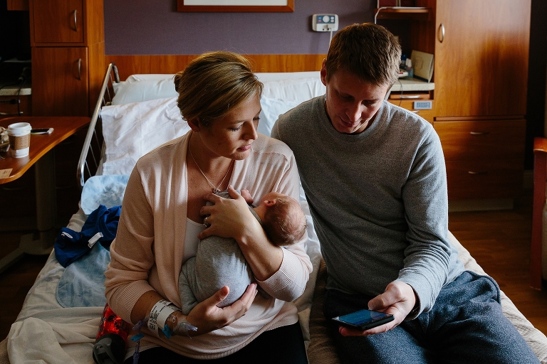 Mother sitting on hospital bed holds newborn baby while father shows weather report on phone