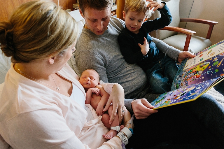 Dad reads a book while mom holds baby boy in the hospital bed.