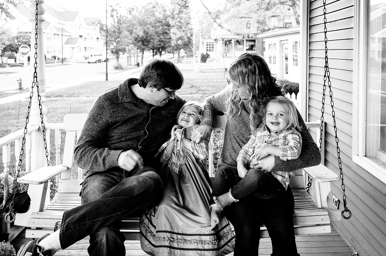 Black and white. Mother, Father, and two daughters all sit on porch swing together