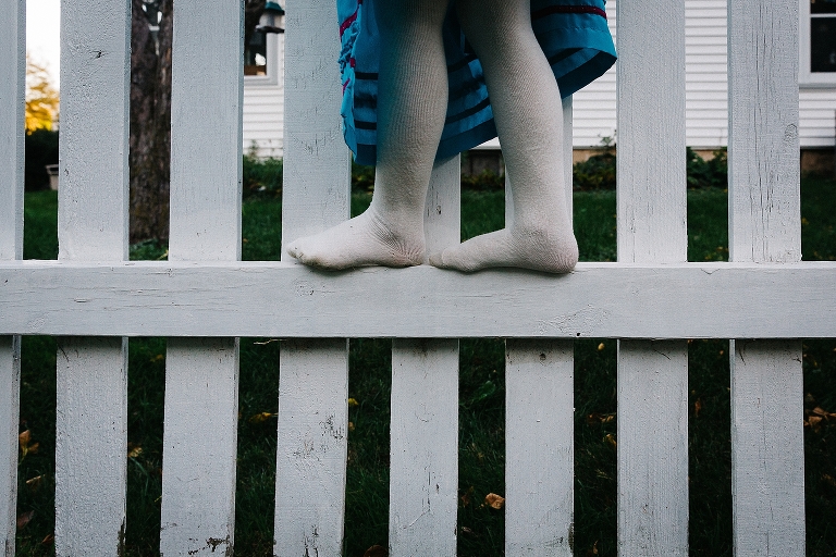 Little girl wearing white tights and no shoes walk on the side of a white fence.