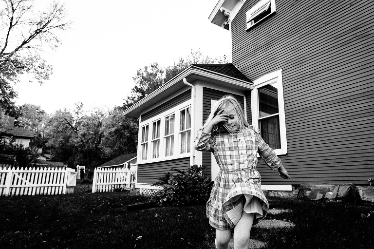 Black and white. Girl wearing plaid dress walks down stone path through her yard