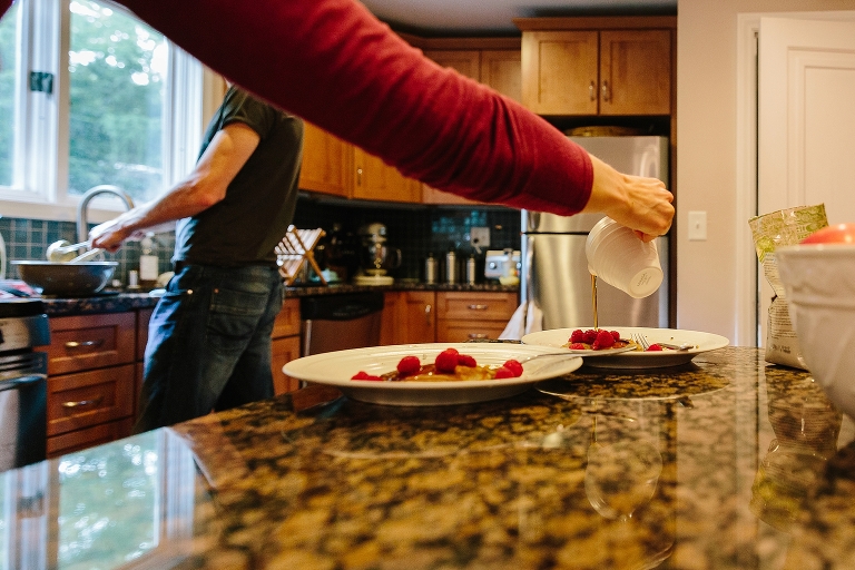 Mom pours syrup on pancakes, Dad ladles pancakes in background 