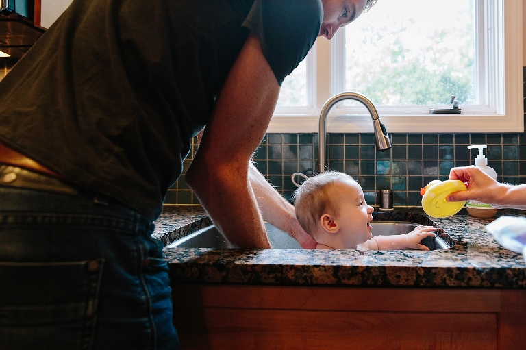 Mom gives daughter a yellow rubber duck in the sink bath while dad holds her