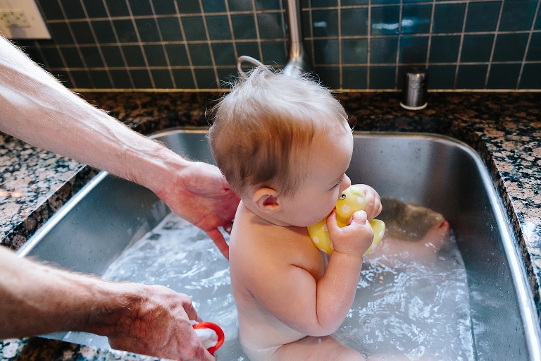 Baby girl chews on yellow duck in sink bath 