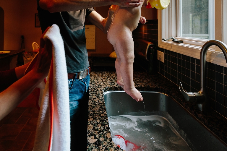Dad holds up baby daughter from a sink bath while mom waits with a towel 