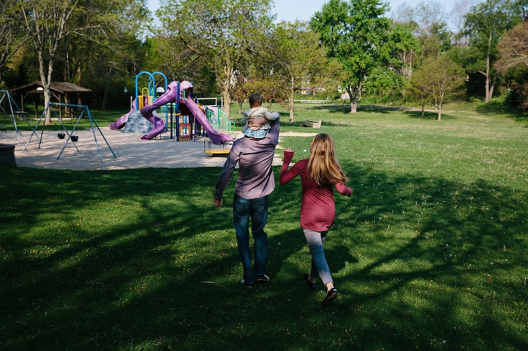 Dad holds daughter on shoulders as he and mom approach a bright playground 