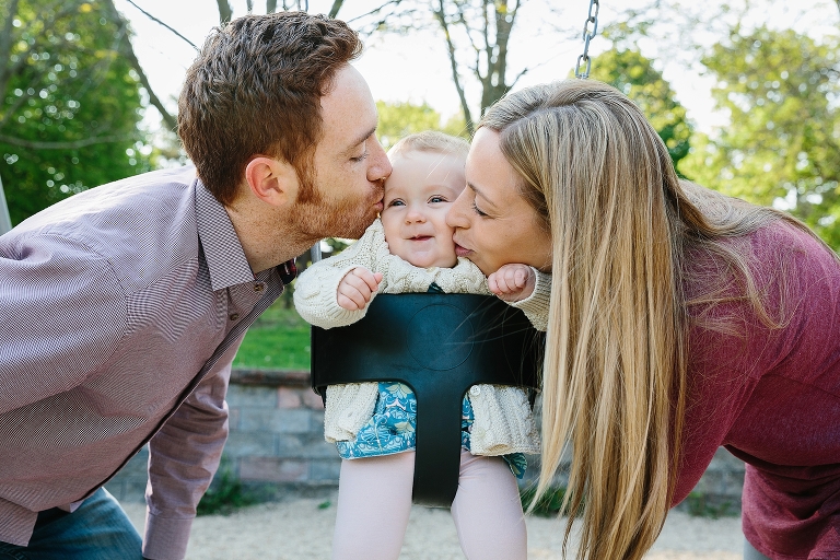 Both Dad and Mom kiss 7 month old daughter on the cheek at the same time while she rides in a swing 