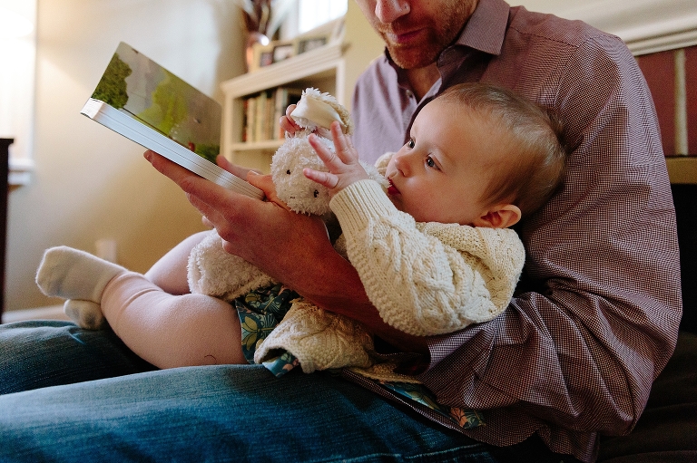 baby girl sits on dad's lap while he reads a book during a Sunday morning pancakes photo session