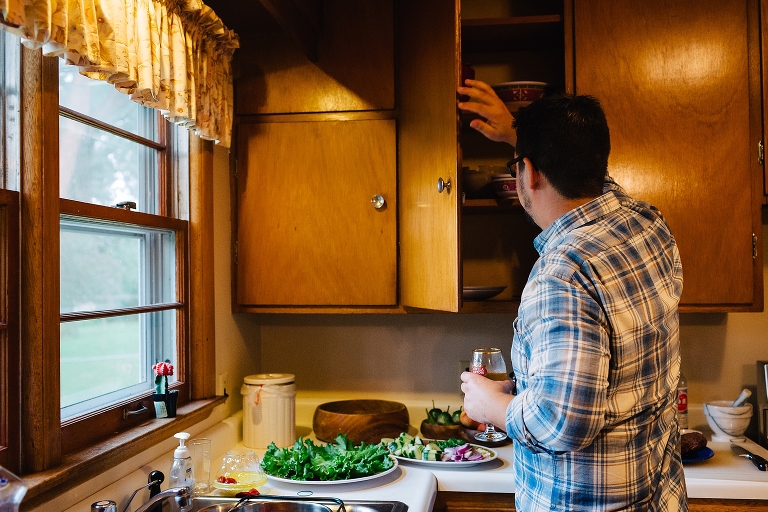 Man in kitchen opening wood cabinet while holding glass of wine
