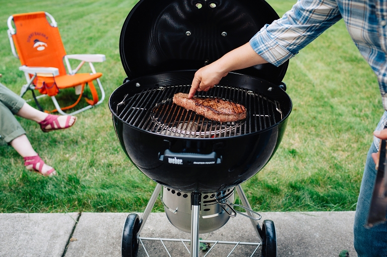 Man cooking piece of red meat outside on grill. lawn chairs laying on grass