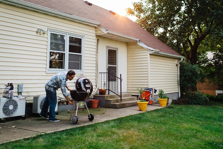 Man outside in backyard patio grilling. Yellow house with sunsetting behind it