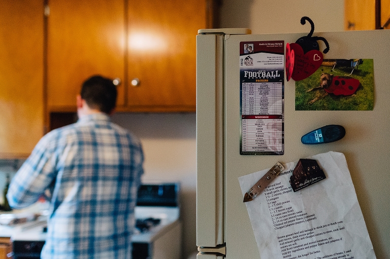 Side of fridge in kitchen with calendar and recipe hanging with magnets. man cooking in background