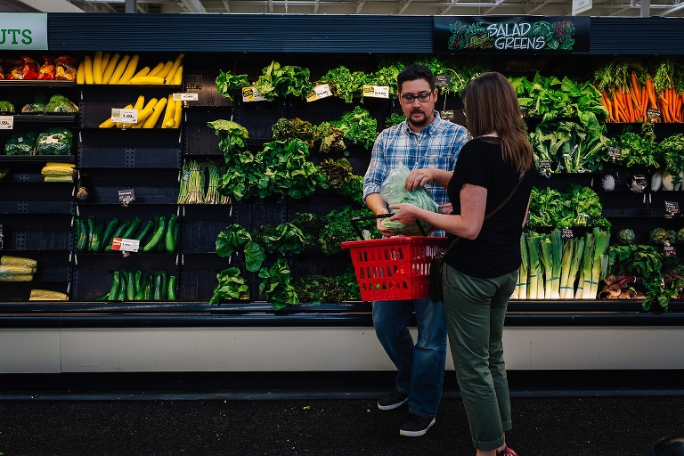 A couple grocery shopping for produce. Man puts bag of lettuce into shopping basket.