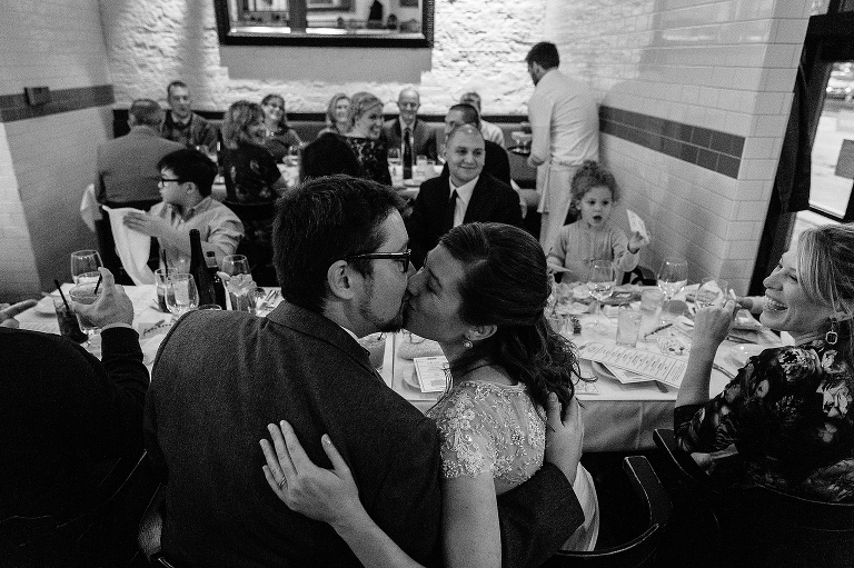 Black and white. Bride and groom kiss at wedding dinner with family.