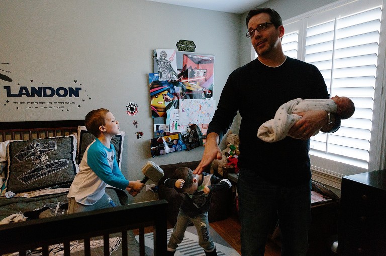 Dad holds newborn daughter while two brother play fight in the background 