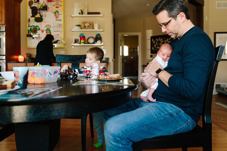 Dad holds newborn daughter at table while son plays with cars. 