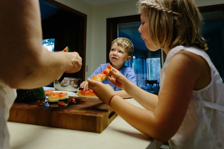 Mom and daughter in foreground, framing young, smiling son. Girl is holding watermelon rind. 