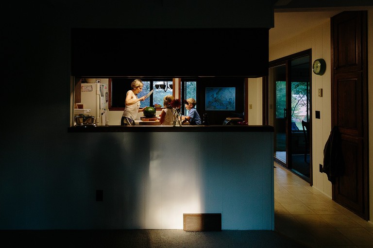 Mom is in mid-slice with a large chef's knife as she is about to cut into a watermelon. Two kids look on. 