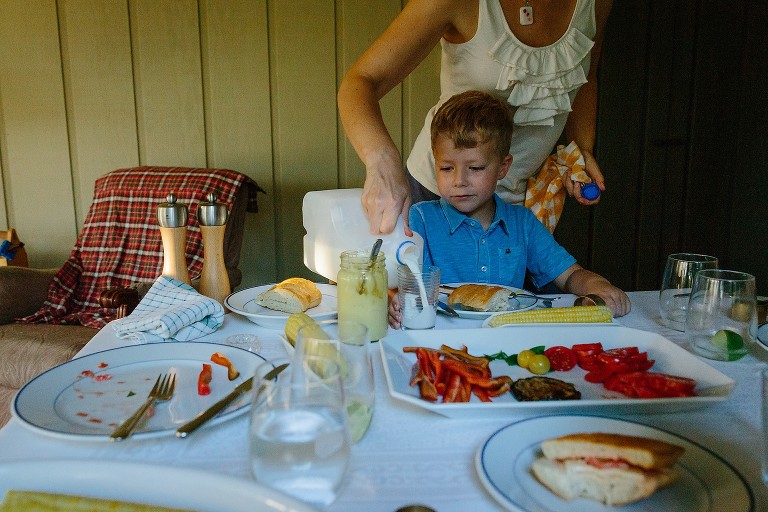 Mom stands behind son and pours a glass of milk. Dinner table has a platter of peppers, eggplant, tomatoes, basil, and cheese.