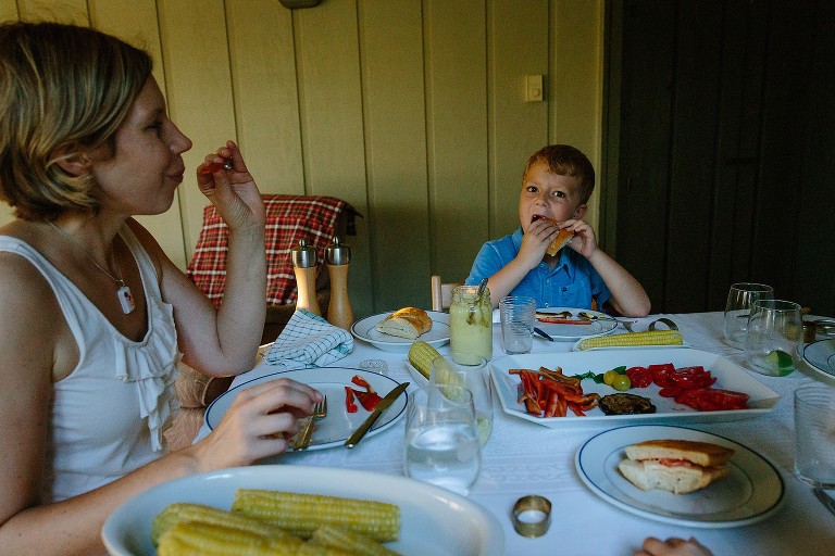 Mom smiles at young boy who is struggling to eat bread without his two front teeth. 