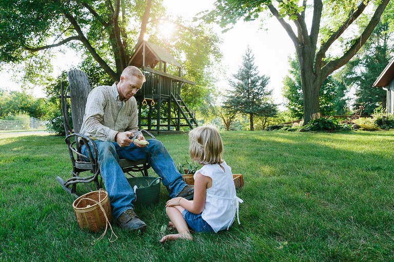 Dad sits in rocking chair and girl sits in grass, sun is shining. They shuck corn.