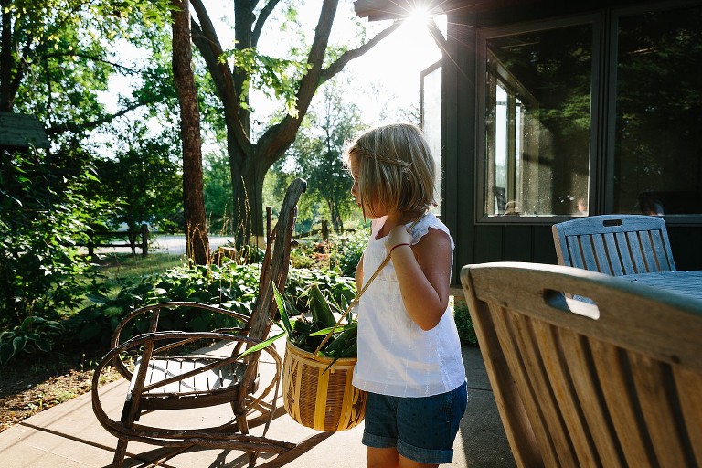 Sun shines on girl holding a basket of corn across shoulder. 