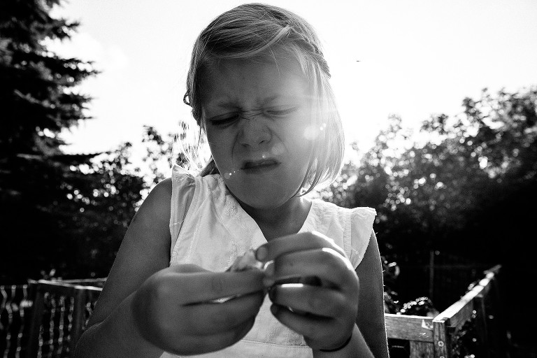 Girl makes a concentrating grimace as she peels a ground cherry. Sun flare enters the frame camera right.
