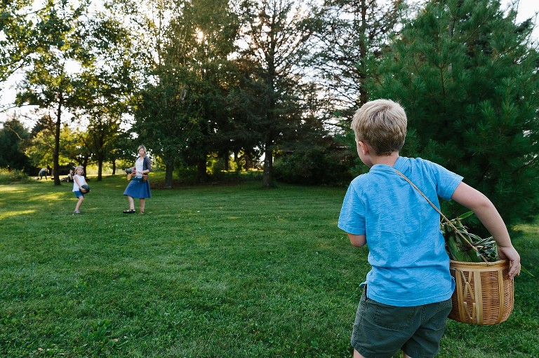 Focus on boy carrying a heavy basket of corn across his shoulders. Mom and sister wait for him 20ft away. 