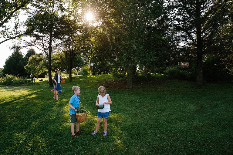 Boy and girl inspect the garden bounty while mom looks toward them. The sun is shining through the trees, the house is in th background. 