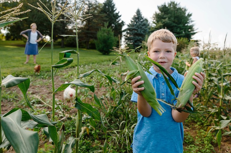 Boy shows off two ears of corn from the garden, Chicken roam the field, mom waits in the background 