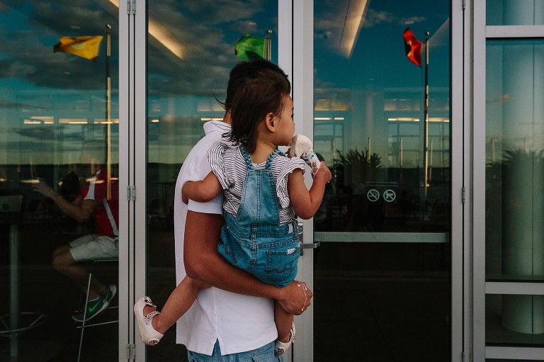 Dad carries daughter, side view, melting  ice cream drips down his hand. Reflection of lake, flags, clouds in window. 