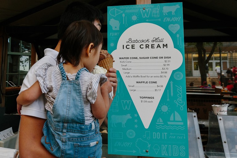 focus on ice cream sign and prices with dad and daughter walking through the frame with cones 