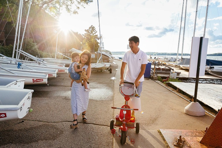 parents walk toddler on path with sailboats and bright sun behind them and the lake to the side 