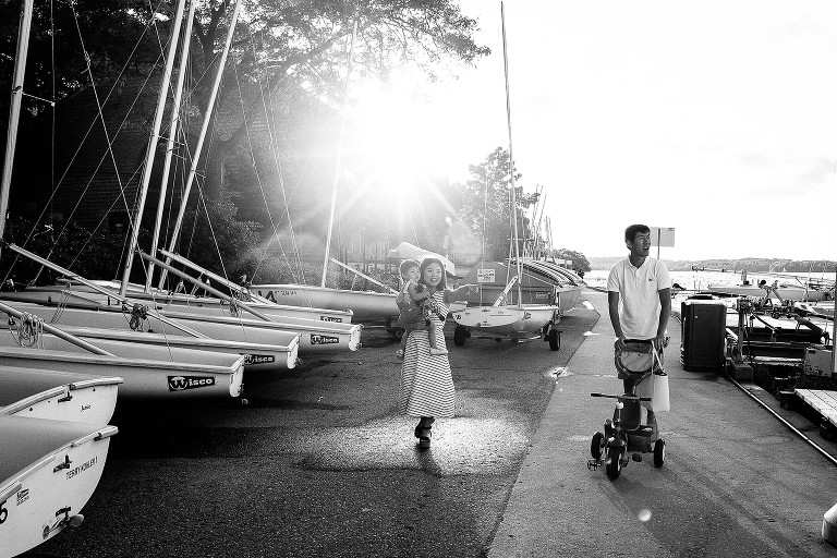 parents walk toddler on path with sailboats and bright sun behind them and the lake to the side 