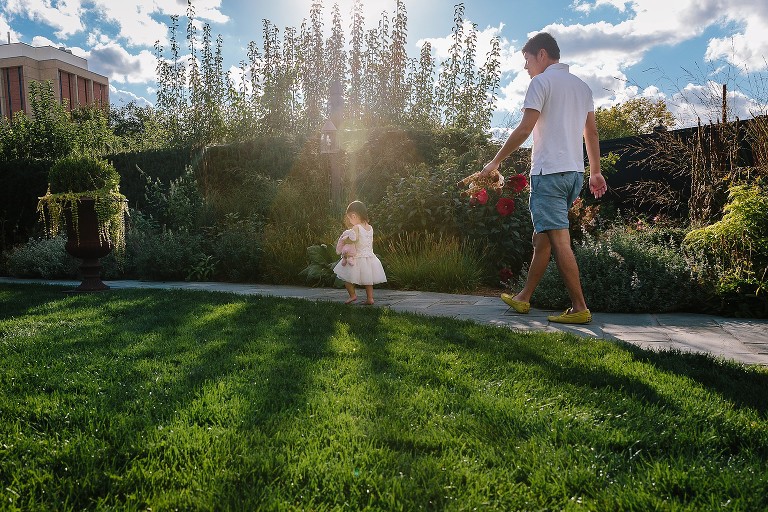 Dad follows young daughter on path. Light and sunflare come through trees