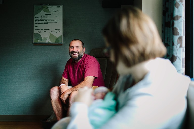 Father smiles at mother and newborn daughter at hte hospital. 