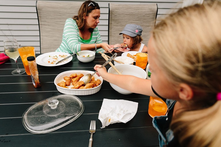 Mom feeds 5 year old dinner, while 7 year old in foreground serves herself orange chicken