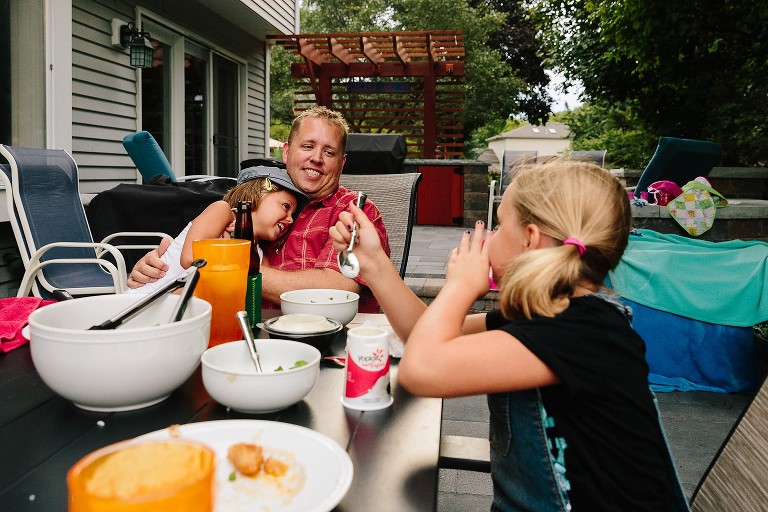 5 year old hugs dad. Dad is laughing and looking at 7 year old telling a joke to him.