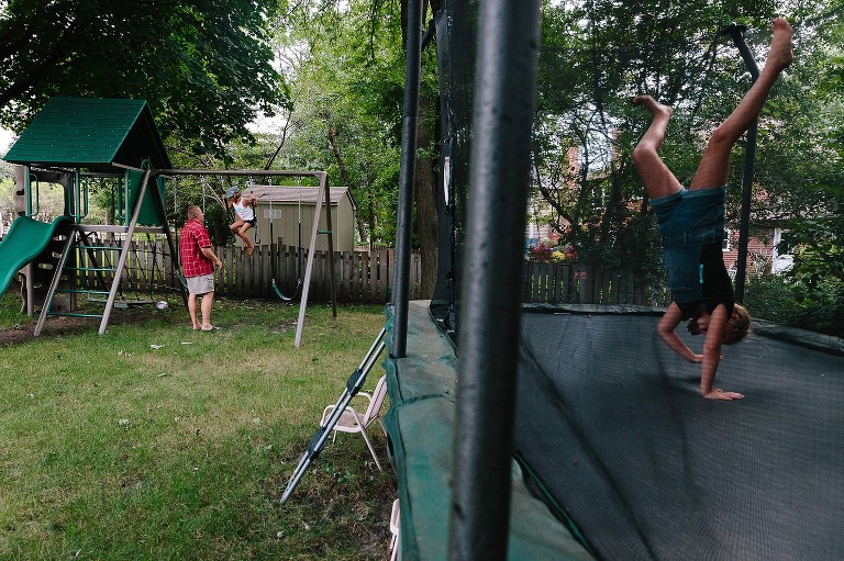 Focus is on Dad and 5yo on swing set, foreground is 7yo doing a cartwheel on trampoline 