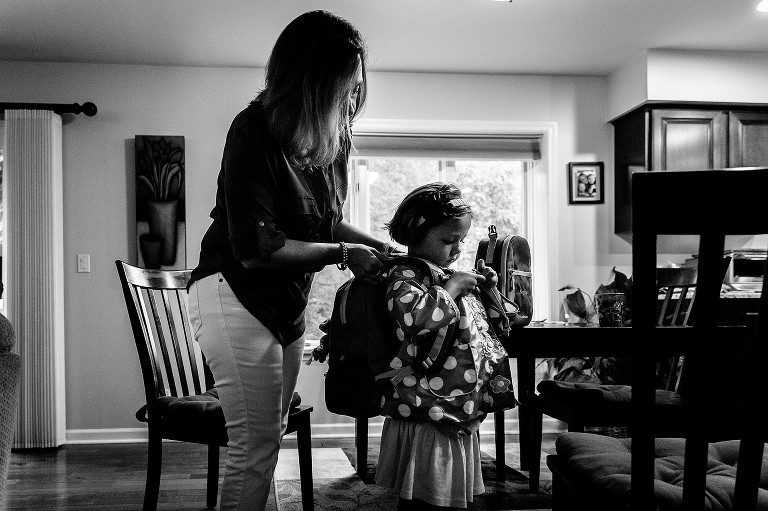 Mom helps daughter get backpack on as she buttons her raincoat 