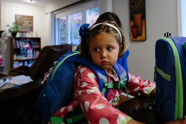 5 year old sits at breakfast table wiht coat and backpack on, nervous expression on her face. 