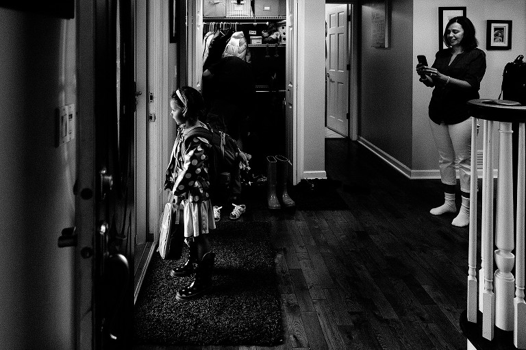 Black and white. Mom uses iPhone to take picture of her daughter standing at the door waiting to leave for school 