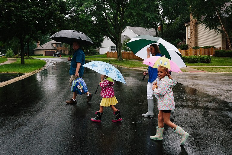 Family crosses neighborhood street in the rain. Everyone has umbrellas. 