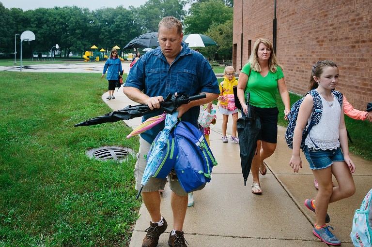 Dad holds all the backpacks and lunchboxes and umbrellas