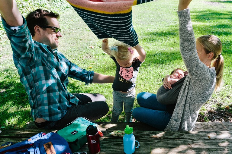 Mom and Dad help daughter take off sweater the park at the same time.
