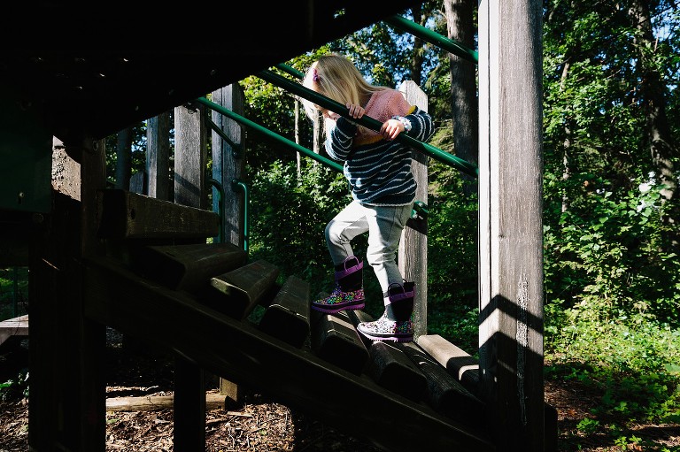 Girl climbs playground equipment in rain boots on a sunny day..