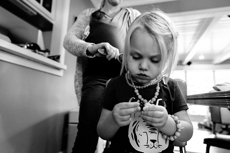 Daughter plays with hair clips while mother brushes hair while wearing sleeping baby