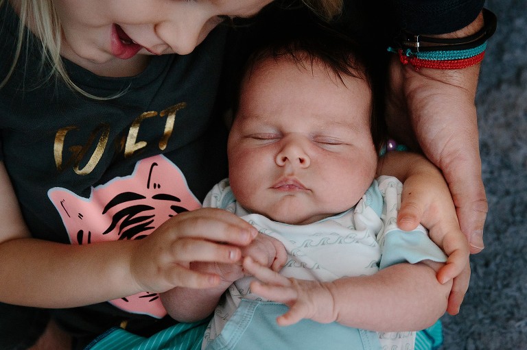Tight shot of sleeping newborn with sister touching his hands, smiling at him.